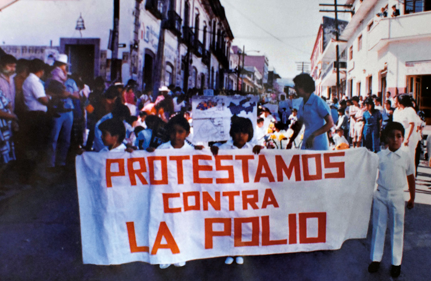 Color photograph of a group of children in white elementary school uniforms marching on a street while a adults watch from the sidewalk. Four children in the front row are holding a large white banner with red letters that read “Protestamos Contra La Polio” (We Protest Against Polio).