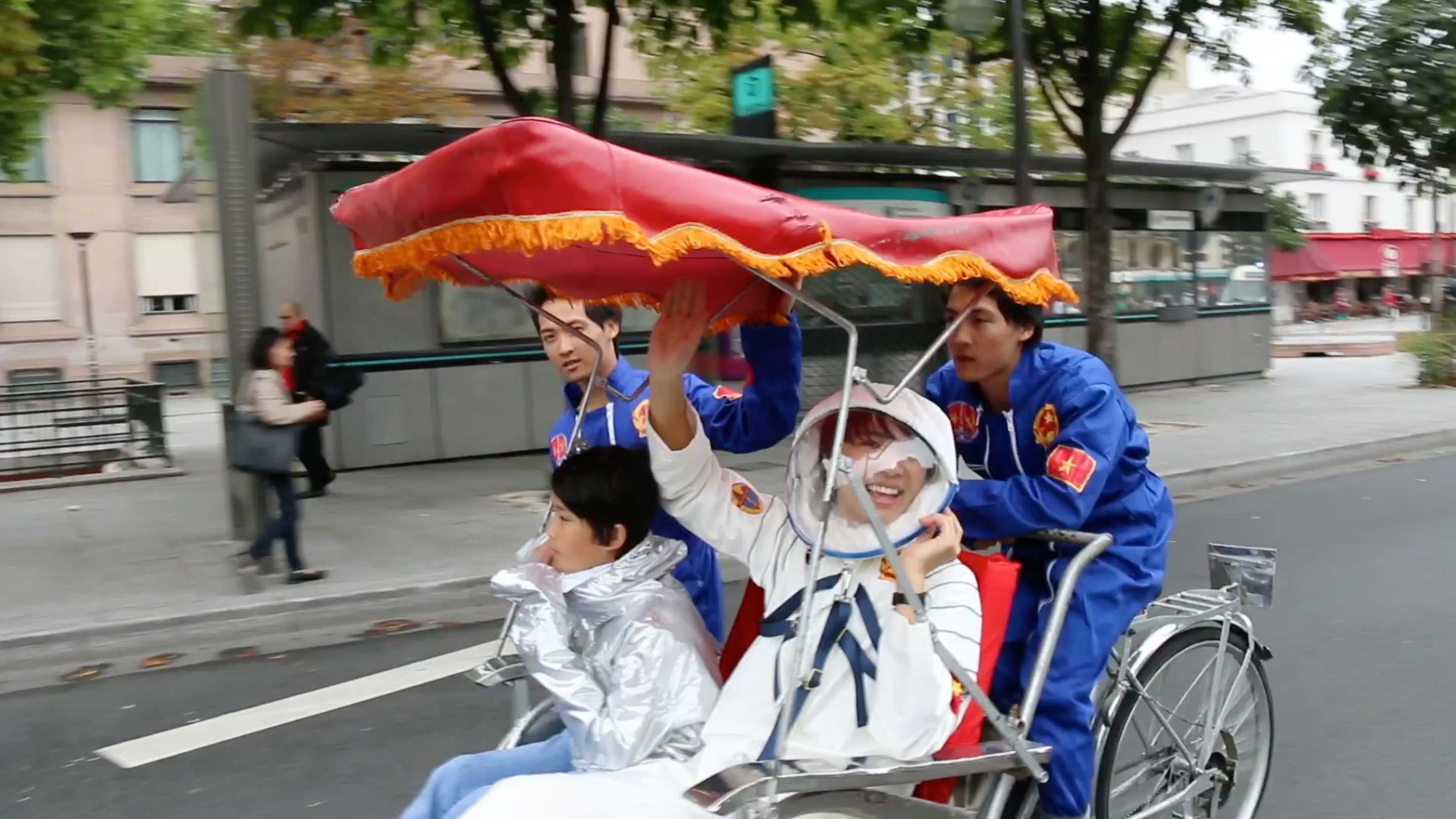 Four people are riding a cyclo through a street. Two who wear blue uniforms are sitting in the back, presumably steering the cyclo, while two who wear a silver space suit and a white astronaut uniform with a helmet sit in the front. The person in the astronaut uniform is laughing and waving.
