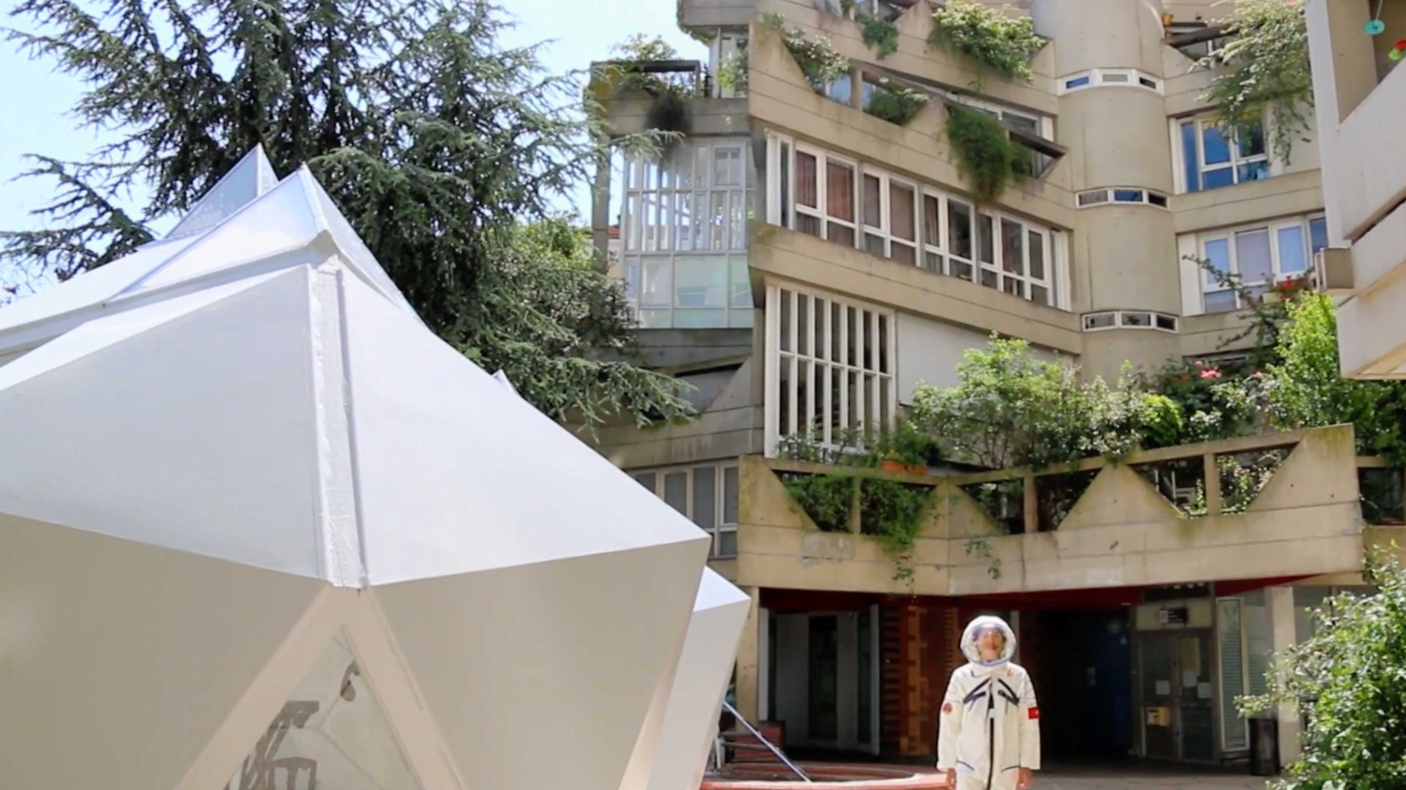 A person in an astronaut uniform stands outside of the angular architecture of Paris’s Ivry-sur-Seine commune. Next to him is a white, inflatable modular structure that matches the star-pointed forms of the housing complex.