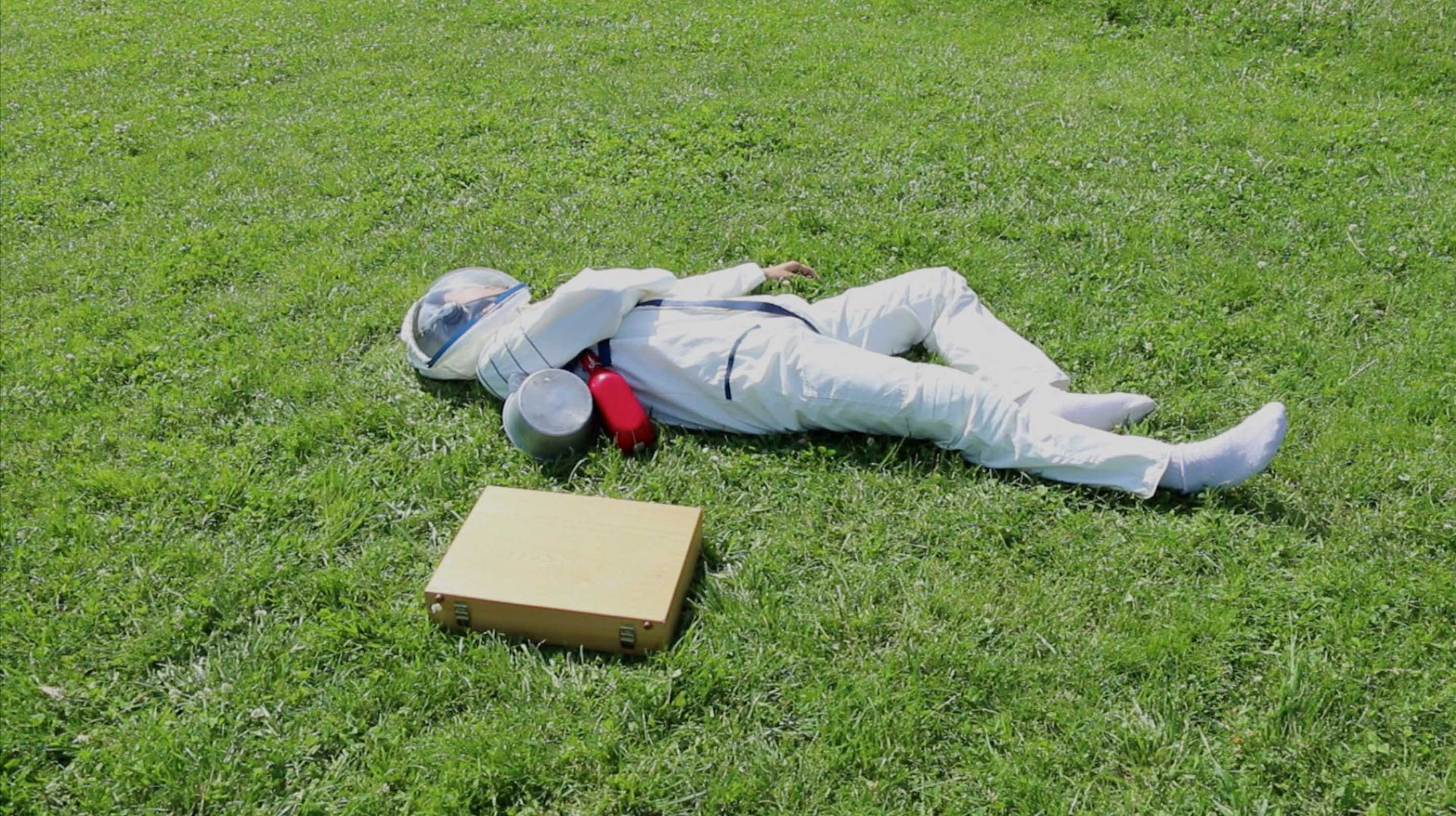 A person in an astronaut uniform with a space helmet lays on the grass next to a golden briefcase.